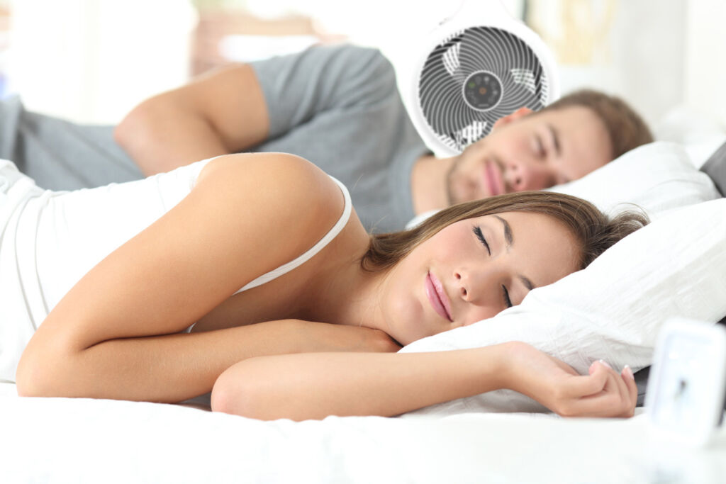 A couple asleep in bed with a fan blowing cool air on them