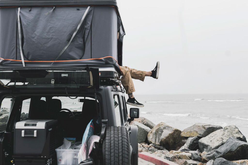 A person sat inside the sleeping area on top of the Jeep camper by the sea