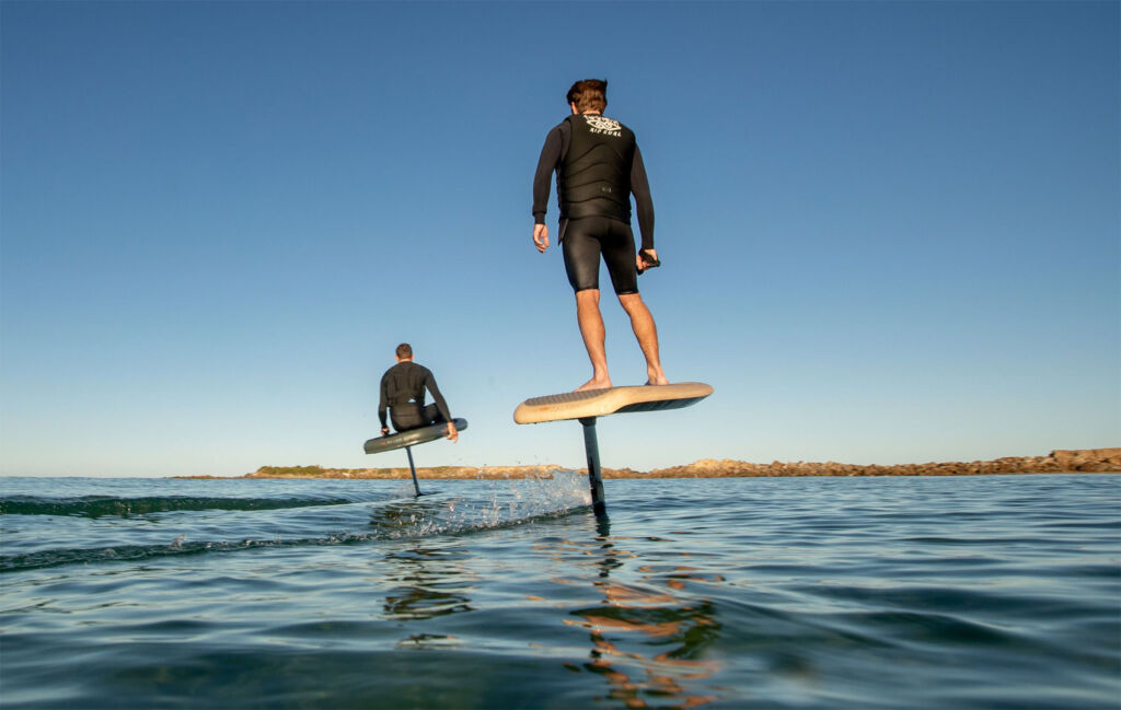Two men using the Fliteboard to travel across a stretch of water, one sitting, one standing.
