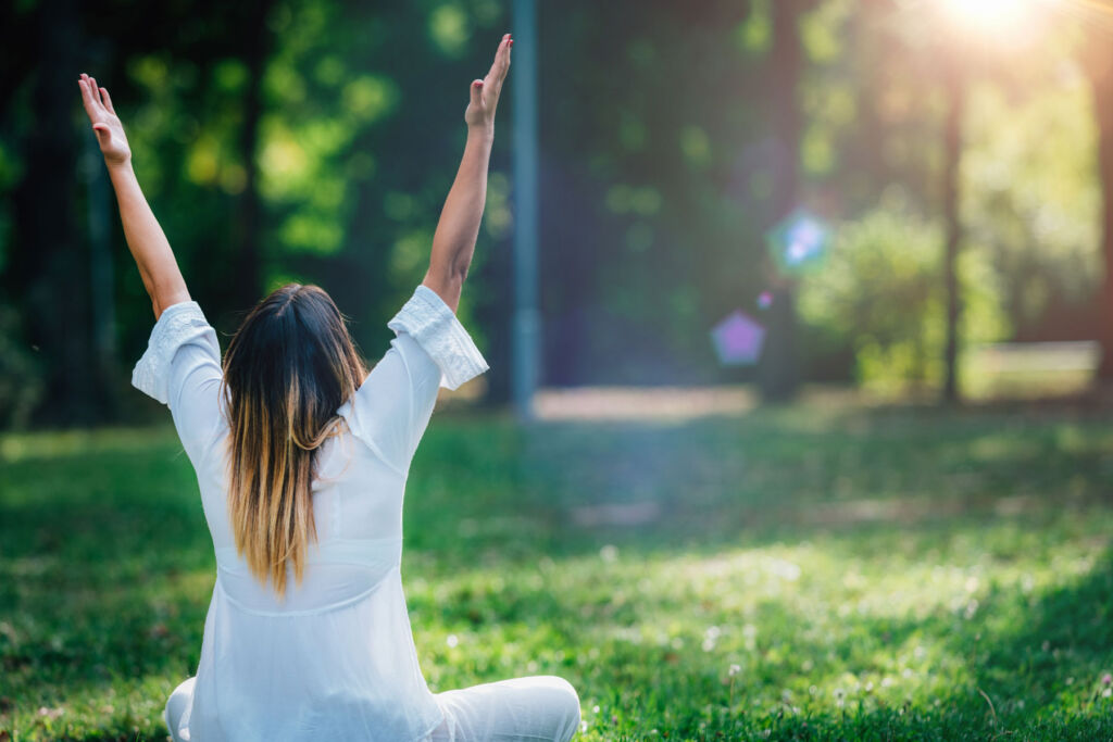A woman raising her hands to the sky whilst sitting on the grass in a park