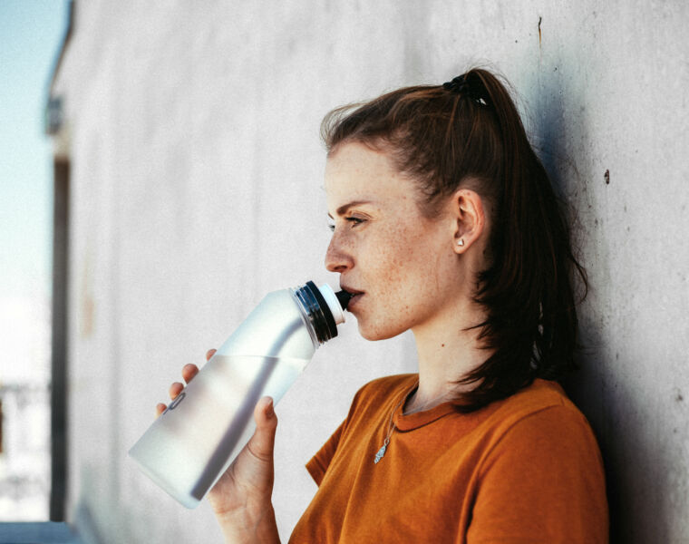 A red head woman leaning against a wall drinking from a bottle