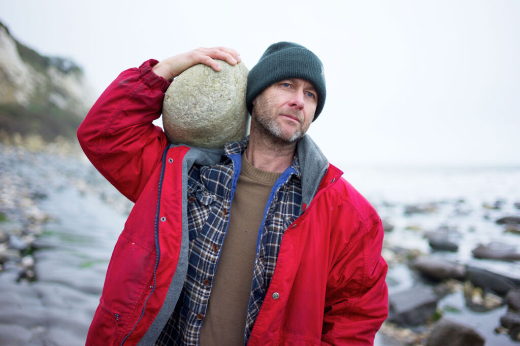 Adrian on a beach with a large boulder on his shoulder
