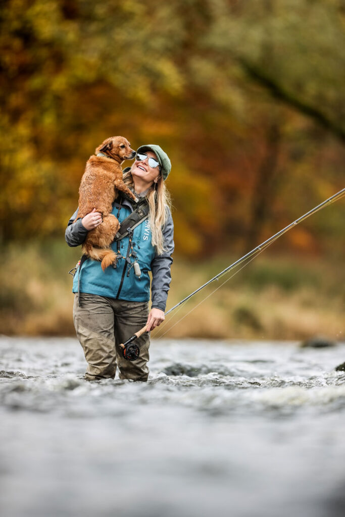 A woman fly fishing in the river whilst holding her dog in her arms