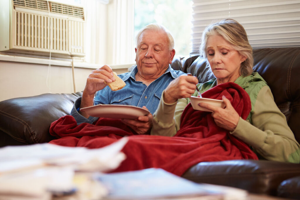 An elderly couple using blankets whilst sat on their sofa to stay warm