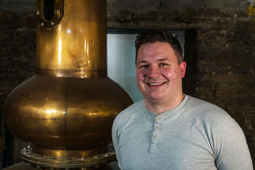 Mark Watson smiling next to one of the distillery stills