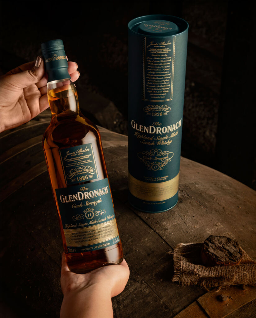A bottle of the new release held in the hand with its box placed on a barrel