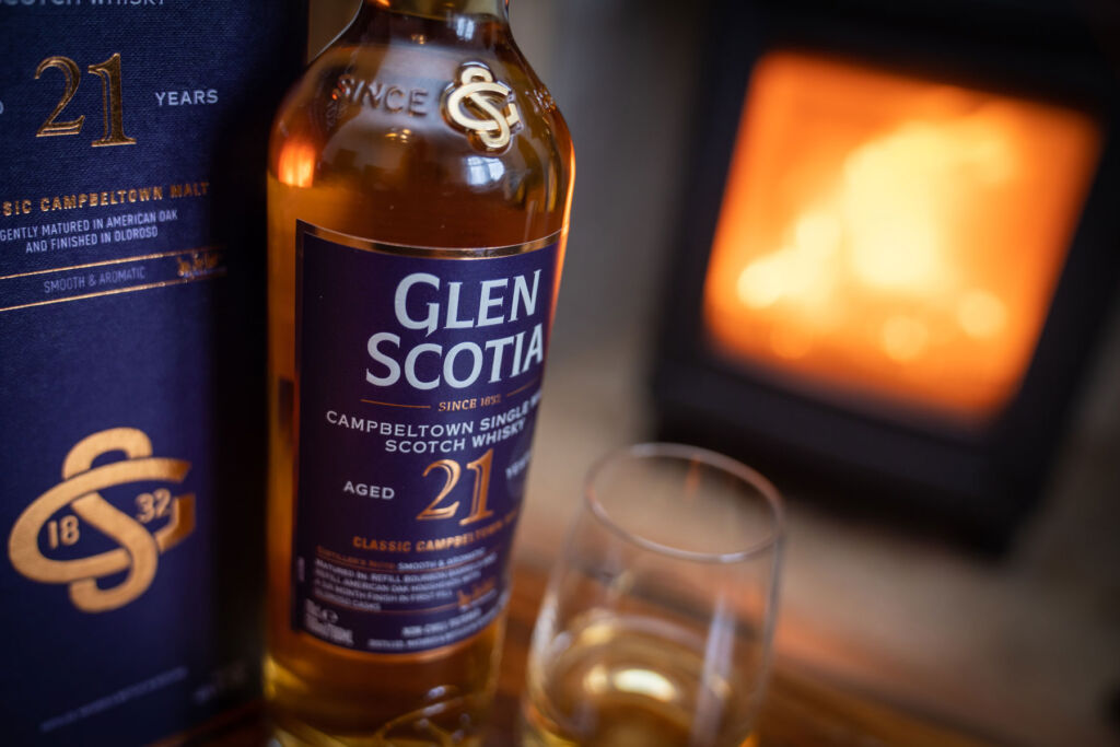 A bottle of the 21-Year-Old Whisky, next to a full glass by a roaring fire