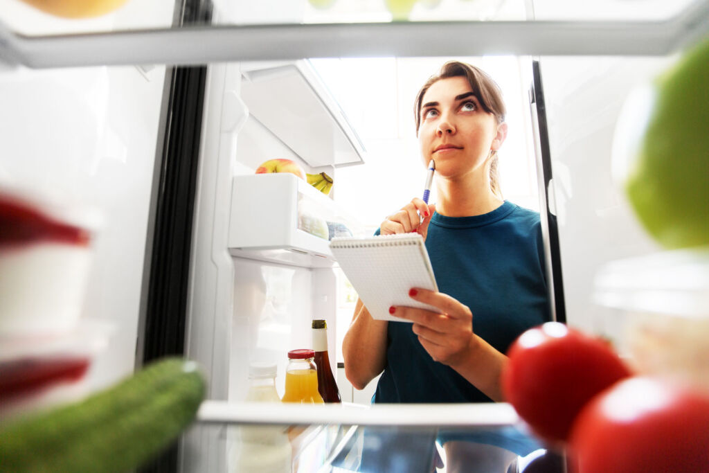 A young woman in front of her fridge writing a list of the foods she needs to get