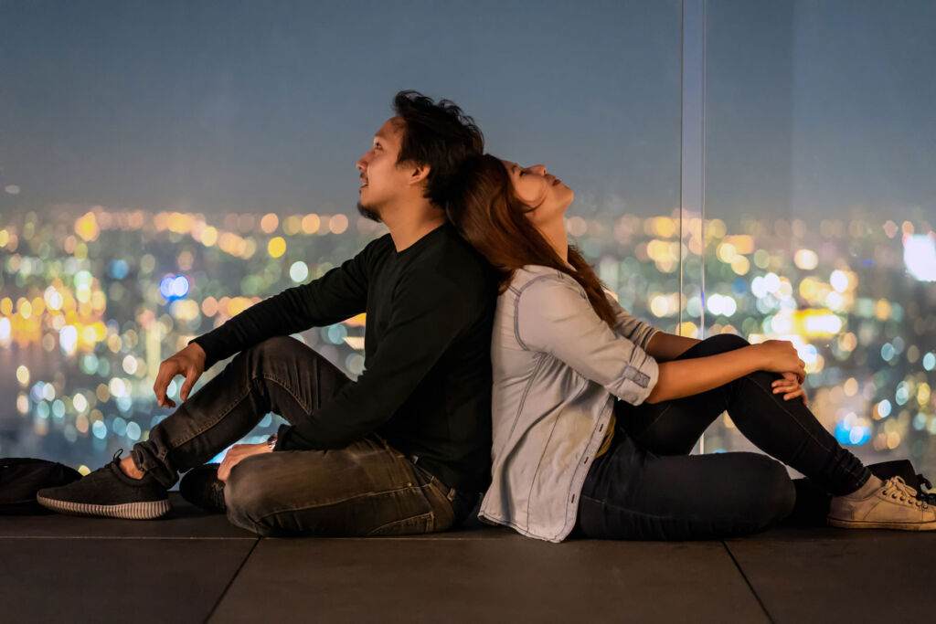 A couple sitting back to back by a window overlooking a city