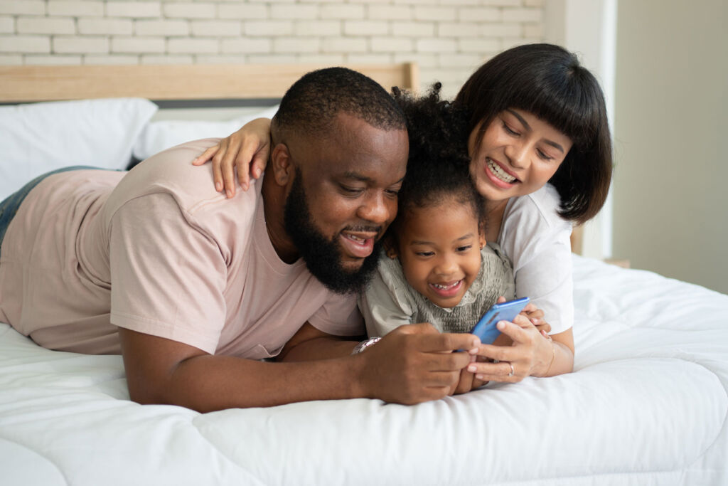 A mother and father on the bed laughing at a mobile phone screen with their young daughter