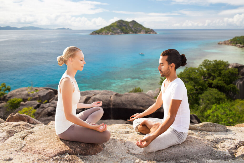 A man and a woman in classic meditation poses atop a cliff by the sea