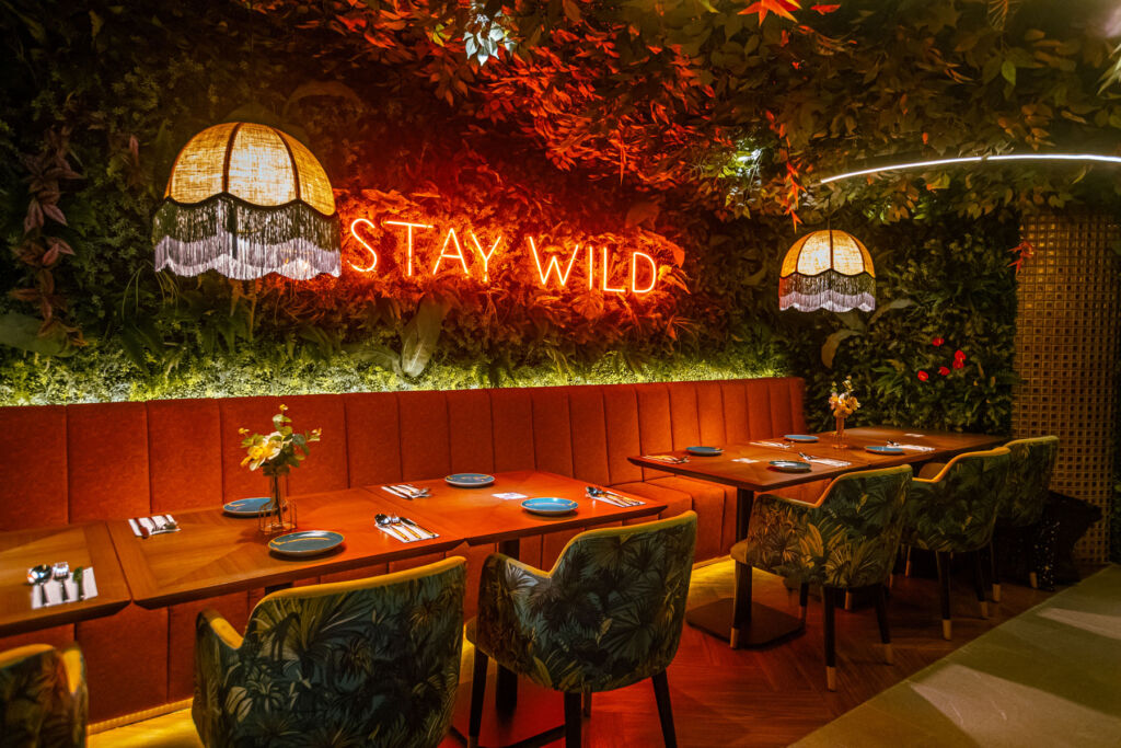 The foliage filled dining are with a neon sign peeking through the leaves with the words, Stay Wild