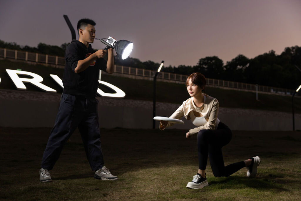 A man lighting a female model, who is throwing a frisbee in the dark