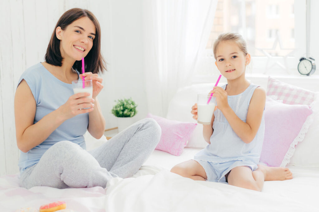 A mother and daughter enjoying a glass of milk whilst sat on a bed