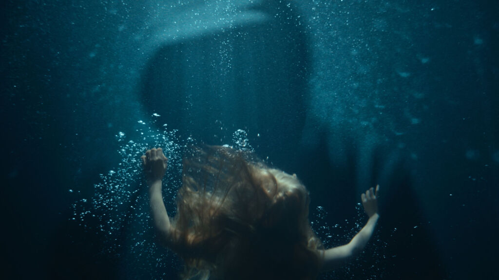 A young girl underwater being faced by a hooded figure