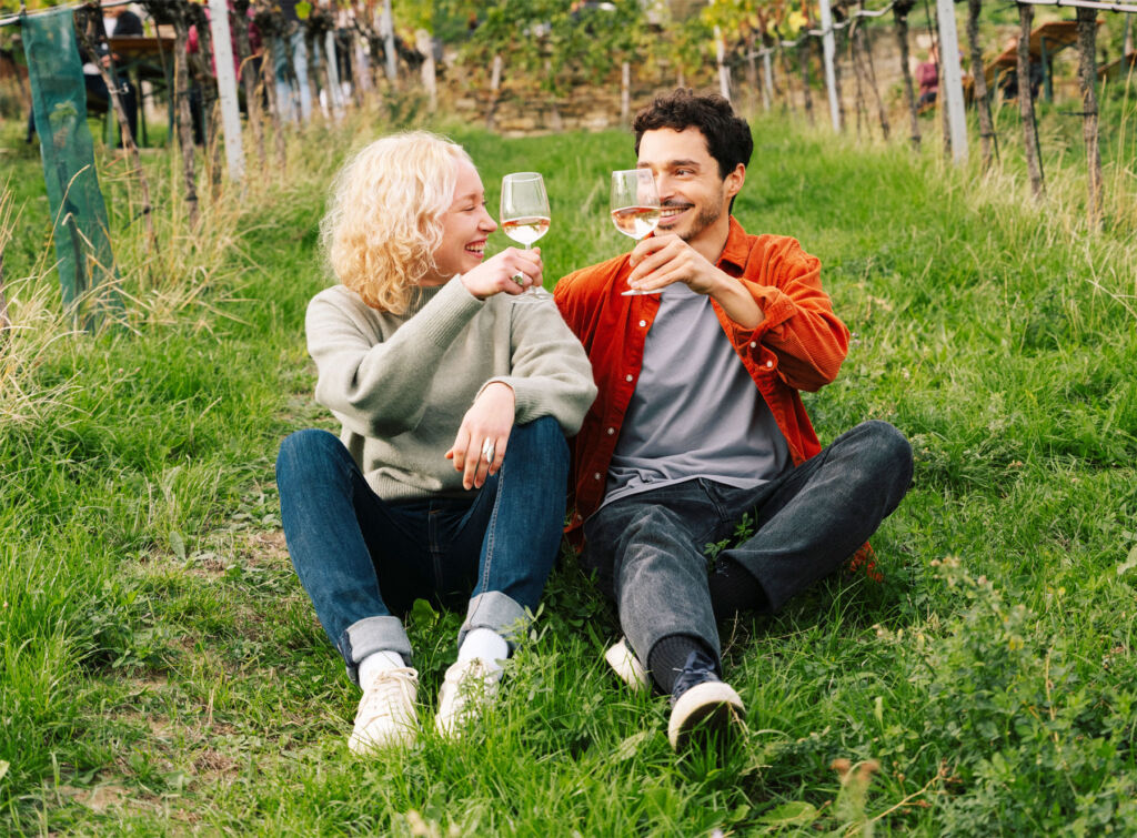 A couple sat on the grass in a vineyard sharing some wine