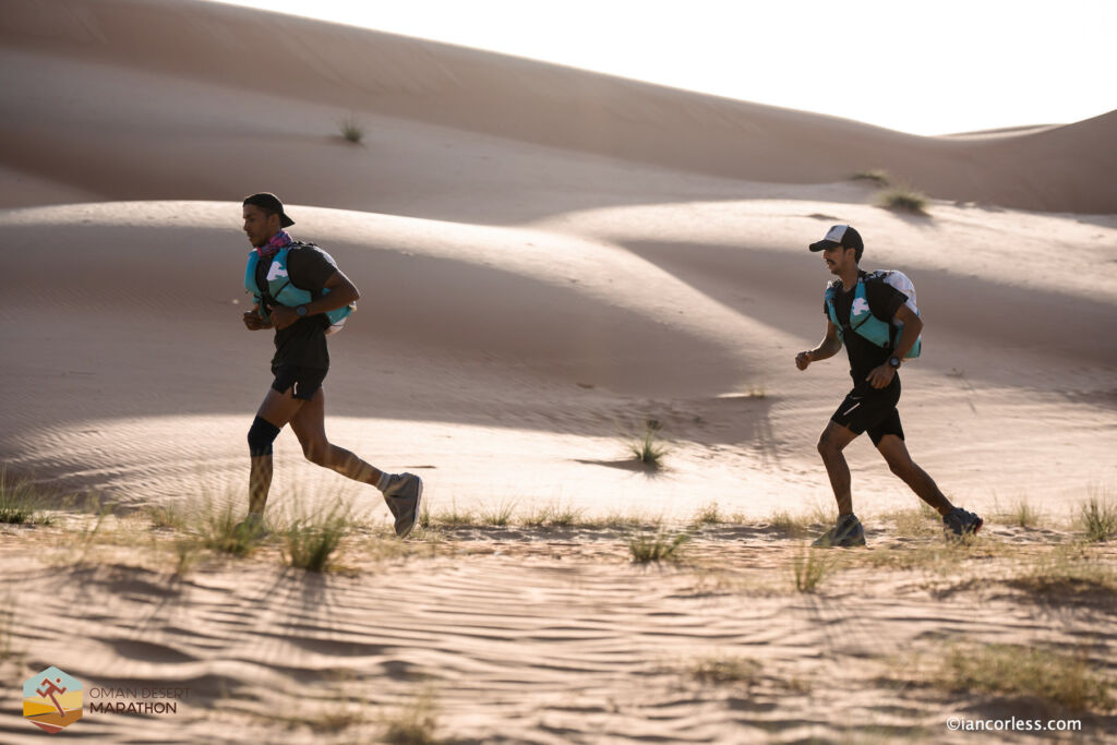 Two male competitors running through the sand as the night falls