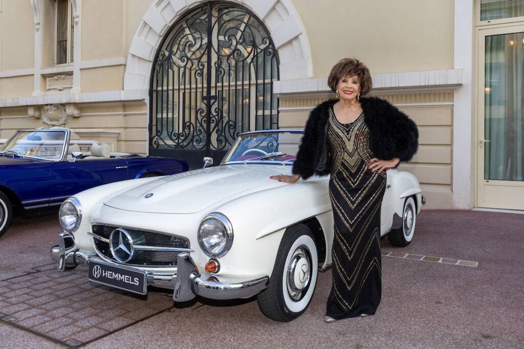 Dame Shirley leaning on the bonnet of her white Mercedes